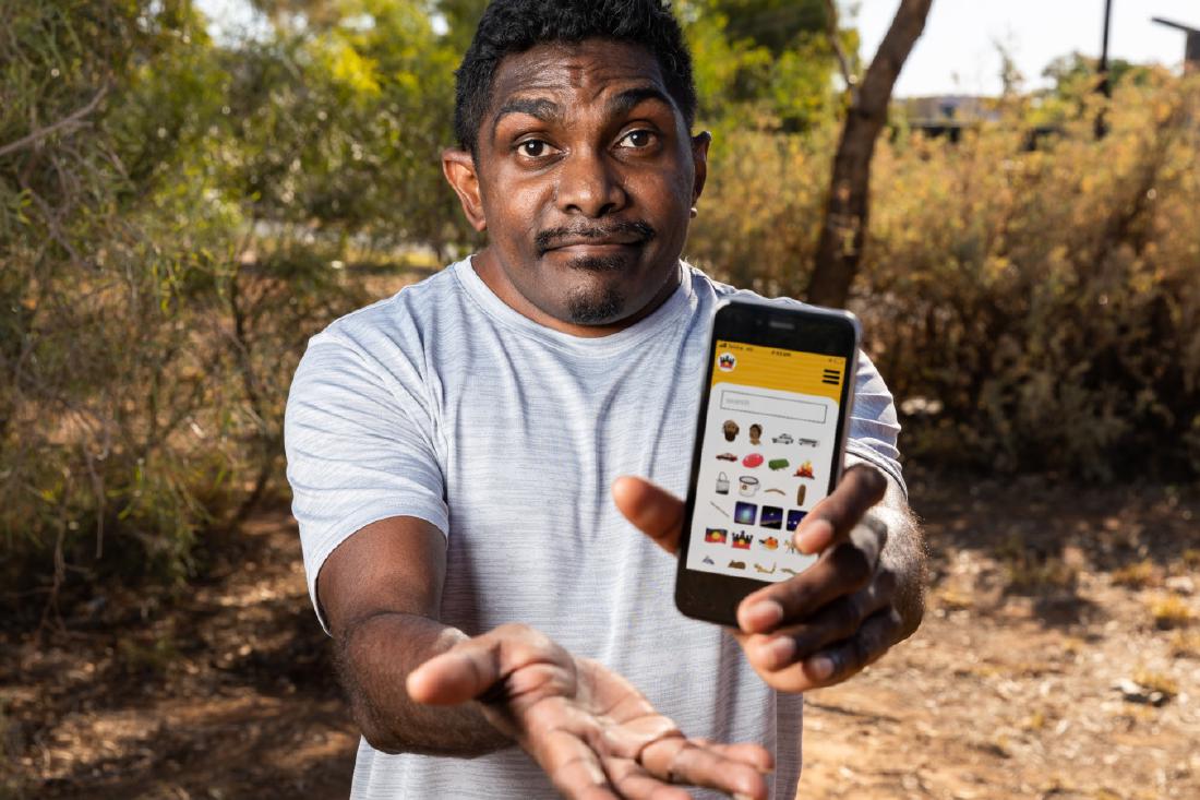 Indigenous person in the forest holds a phone displaying the Indigemoji application on it.